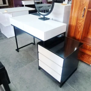 میز آرایش زیبا و با کیفیت مدل لیندا AVI-DRT035 میز آرایش مدل لیندا AVI-DRT035 با رنگ چوب طبیعی و فرم هندسی ساده، جلوه‌ای مدرن و مینیمال به فضای اتاق خواب می‌بخشد. این محصول از ام‌دی‌اف با کیفیت بالا ساخته شده و سطح آن با پوشش ضدخش و مقاوم در برابر رطوبت تکمیل شده است. طراحی دو کشوی افقی در بخش میز و فایل سه‌کشوی جانبی، فضای کافی برای نگهداری لوازم آرایشی، زیورآلات و وسایل شخصی فراهم می‌کند. پایه‌ها از پروفیل فلزی سبک با رنگ الکترواستاتیک مشکی مات ساخته شده‌اند که علاوه بر استحکام، زیبایی صنعتی خاصی به محصول می‌دهند. طراحی بدون دستگیره کشوها این میز آرایش، ظاهر یکپارچه و مدرنی ایجاد کرده است. کیفیت ساخت در ساخت میز آرایش مدل لیندا AVI-DRT035 ، دقت در جزئیات و انتخاب متریال باکیفیت نقش مهمی ایفا کرده است. کشوها با ریل‌های فلزی نرم‌بازشو تجهیز شده‌اند که عملکرد روان و بی‌صدا را تضمین می‌کنند. اتصالات داخلی محصول آرایش مدل لیندا با پیچ‌های مخفی و تقویت‌شده طراحی شده‌اند تا دوام و پایداری محصول در طول زمان حفظ شود. رنگ چوب گرم و طبیعی، حس آرامش و هماهنگی را به محیط منتقل می‌کند و با پایه‌های فلزی مشکی مات، تضاد بصری جذابی ایجاد می‌شود. این میز آرایش نه‌ تنها کاربردی است بلکه به‌عنوان یک عنصر دکوراتیو نیز عمل می‌کند.  زیبایی‌شناسی و عملکرد این محصول ترکیب متریال با کیفیت و طراحی مدرن، میز آرایش مدل لیندا AVI-DRT035 را به گزینه‌ای خاص برای دکوراسیون داخلی تبدیل کرده است. فرم هندسی ساده، خطوط تمیز و رنگ‌بندی متعادل، جلوه‌ای حرفه‌ای و شیک به محصول بخشیده‌اند. این میز نه‌تنها فضای ذخیره‌سازی کافی برای لوازم آرایشی و شخصی فراهم می‌کند، بلکه با ظاهر چشم‌نواز خود، نقطه کانونی اتاق خواب خواهد بود.  کیفیت ساخت بالا، مقاومت MDF در برابر رطوبت و فشار، و استقامت پایه‌های فلزی با رنگ ثابت از ویژگی‌های برجسته این محصول‌اند. آینه مدور نصب‌شده روی میز، زیبایی و کارایی را هم‌زمان ارائه می‌دهد. این میز آرایش انتخابی عالی برای کسانی است که به طراحی حرفه‌ای، فضای سازمان‌یافته و زیبایی بصری اهمیت می‌دهند.